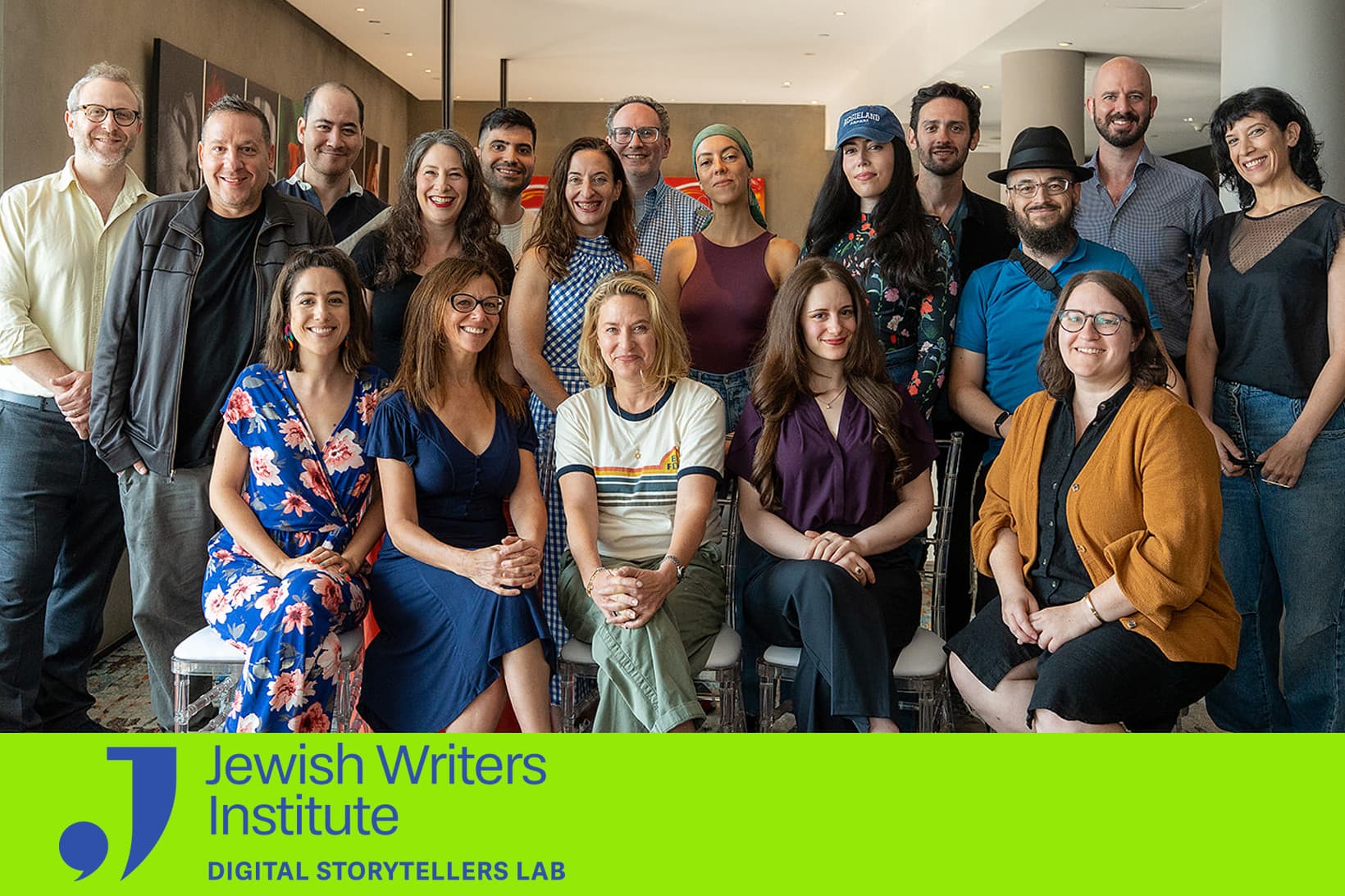 A group portrait of sixteen diverse individuals associated with the Jewish Writers Institute Digital Storytellers Lab. The group is arranged in two rows—five people are seated in the front, and eleven are standing behind them—inside a brightly lit, modern indoor space with art on the walls. They are dressed in casual to smart-casual attire and are smiling at the camera. At the bottom of the image, a vibrant lime-green banner features the organization's logo: a stylized blue letter "J" that incorporates a comma-like shape, followed by the text "Jewish Writers Institute" and "DIGITAL STORYTELLERS LAB" in blue sans-serif font.