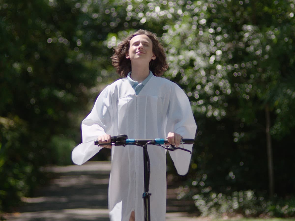 A medium shot of Ethan Bloom, a young man with shoulder-length wavy brown hair, riding an electric scooter down a sun-dappled, tree-lined path. He wears a flowing white graduation gown over a light blue polo shirt. His eyes are closed and his head is tilted back slightly toward the sky with a serene, blissful expression. The background is a lush, green forest with soft, circular bokeh patterns created by sunlight filtering through the leaves.