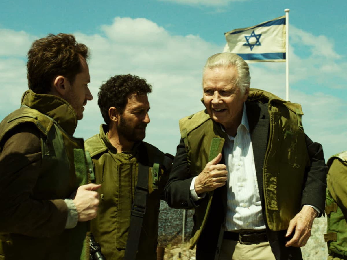 A wide film still showing an older, light-skinned man with swept-back white hairâresembling actor Jon Voightâsmiling broadly as he is helped into a green military tactical vest. He wears a light, striped dress shirt, khaki trousers, and a dark blazer underneath the vest. He is surrounded by four younger men dressed in olive-green military uniforms. To his left, a man with dark curly hair and a beard assists him. To his right, another soldier with sunglasses watches on. In the background, an Israeli flag flies against a bright blue sky with scattered white clouds, and elements of a military outpost or checkpoint are visible. The overall color grading has a distinct warm, greenish-yellow tint.