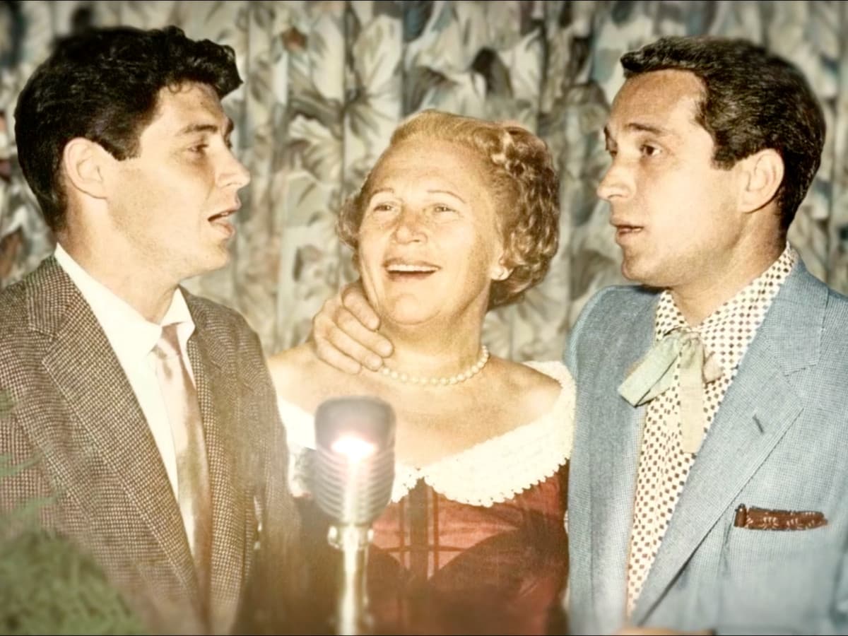 A vintage-style, colorized photograph featuring three people gathered around a classic silver microphone. In the center, a woman with blonde hair and a pearl necklace smiles warmly. She is flanked by two young men in suits; the man on the left appears to be singing or speaking into the microphone, while the man on the right looks on. The background consists of floral-patterned curtains, evoking a mid-20th-century resort atmosphere.